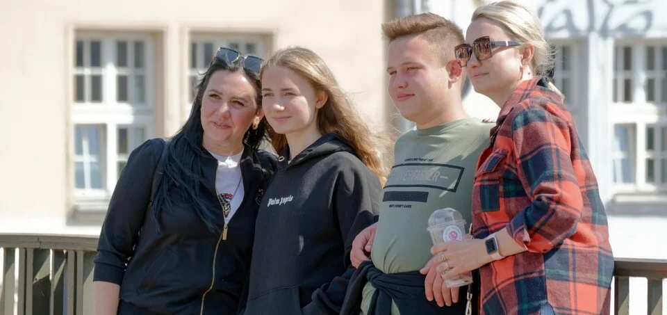 Eine Gruppe junger Leute stehen an einem Geländer und sehen in die selbe Richtung. Eine Frau mit Sonnenbrille hat einen Kaffeebescher in der Hand.